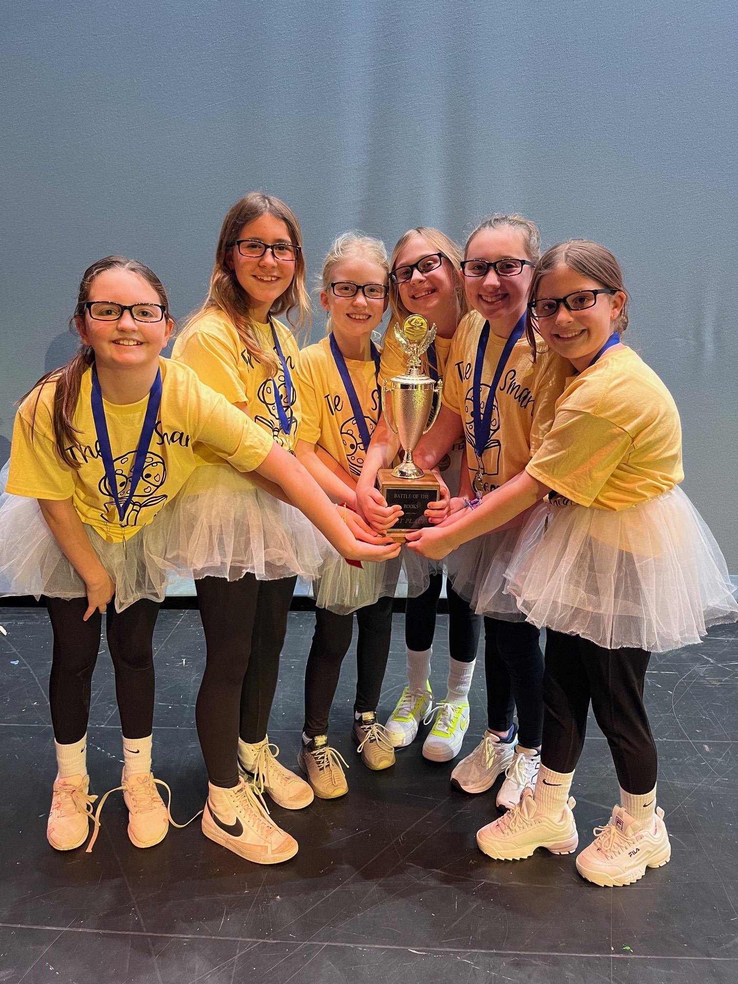 Championship team holding trophy. Six girls in yellow shirts and white tutus smiling with pride.
