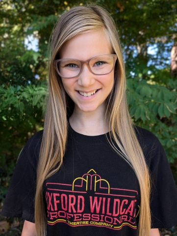 Young lady with long blonde hair and glasses wearing a black T-shirt with Oxford Wildcat Theatre Company written on it.